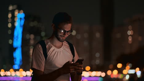 A-male-traveler-with-a-backpack-with-glasses-on-the-background-of-the-night-city-calls-a-taxi-through-a-mobile-phone.-Holds-a-smartphone-and-touches-the-screen.-See-photos-and-write-messages-in-social-networks